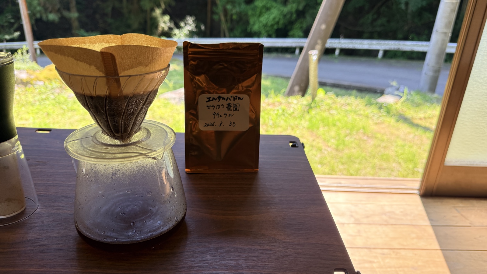 山の家の窓辺で、エルサルバドル・マラカウ農園のコーヒーをドリップで淹れている様子。窓の外には緑の山が広がる