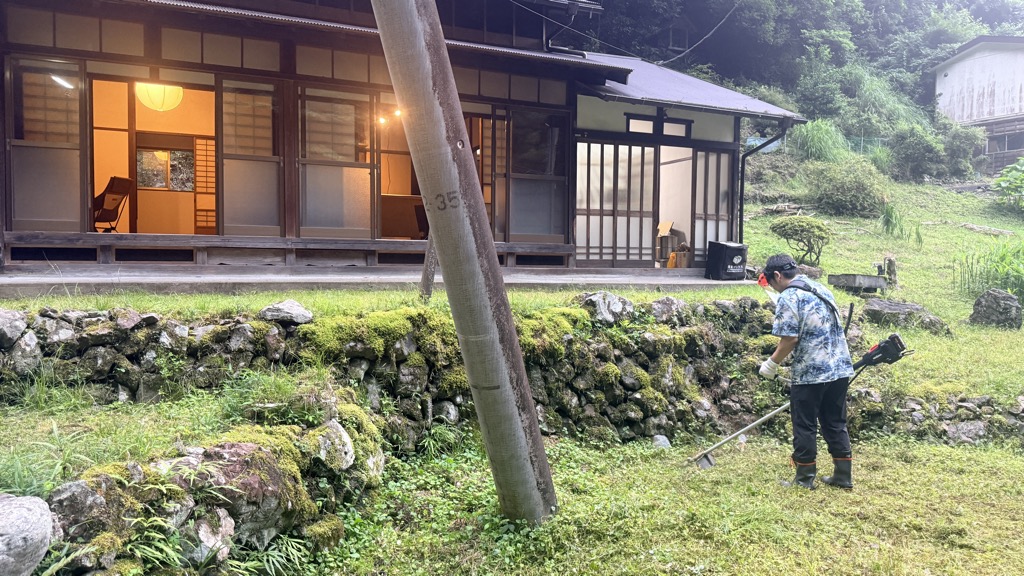 山の家の庭で夕方にヘッドライトを付けて草刈り機を使う協力者の写真